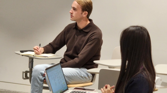 Man sitting down looking in the distance at a desk with a pen and paper. Girl in the forefront also sitting down, looking at the man.