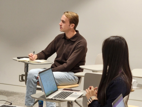 Man sitting down looking in the distance at a desk with a pen and paper. Girl in the forefront also sitting down, looking at the man.